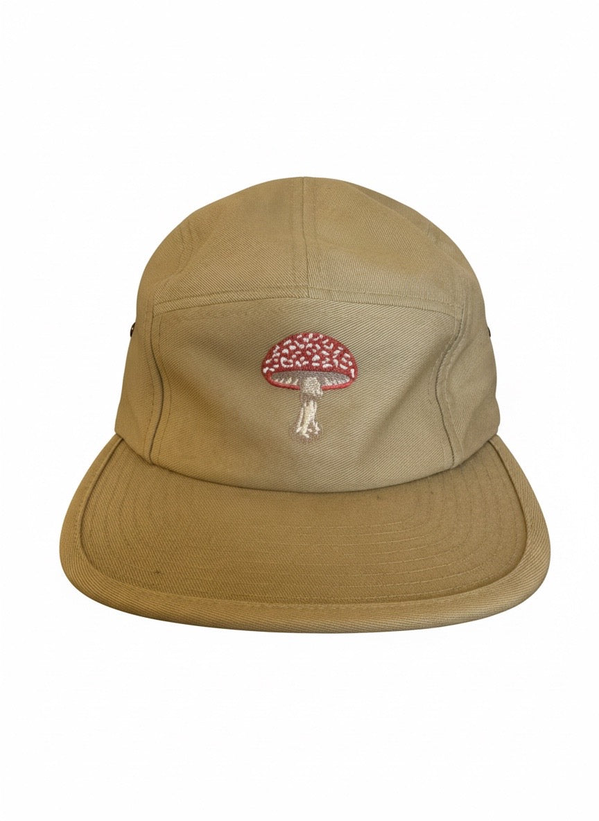 A tan 5-panel camp cap with a small embroidered red and white mushroom patch on the front, set against a plain white background.